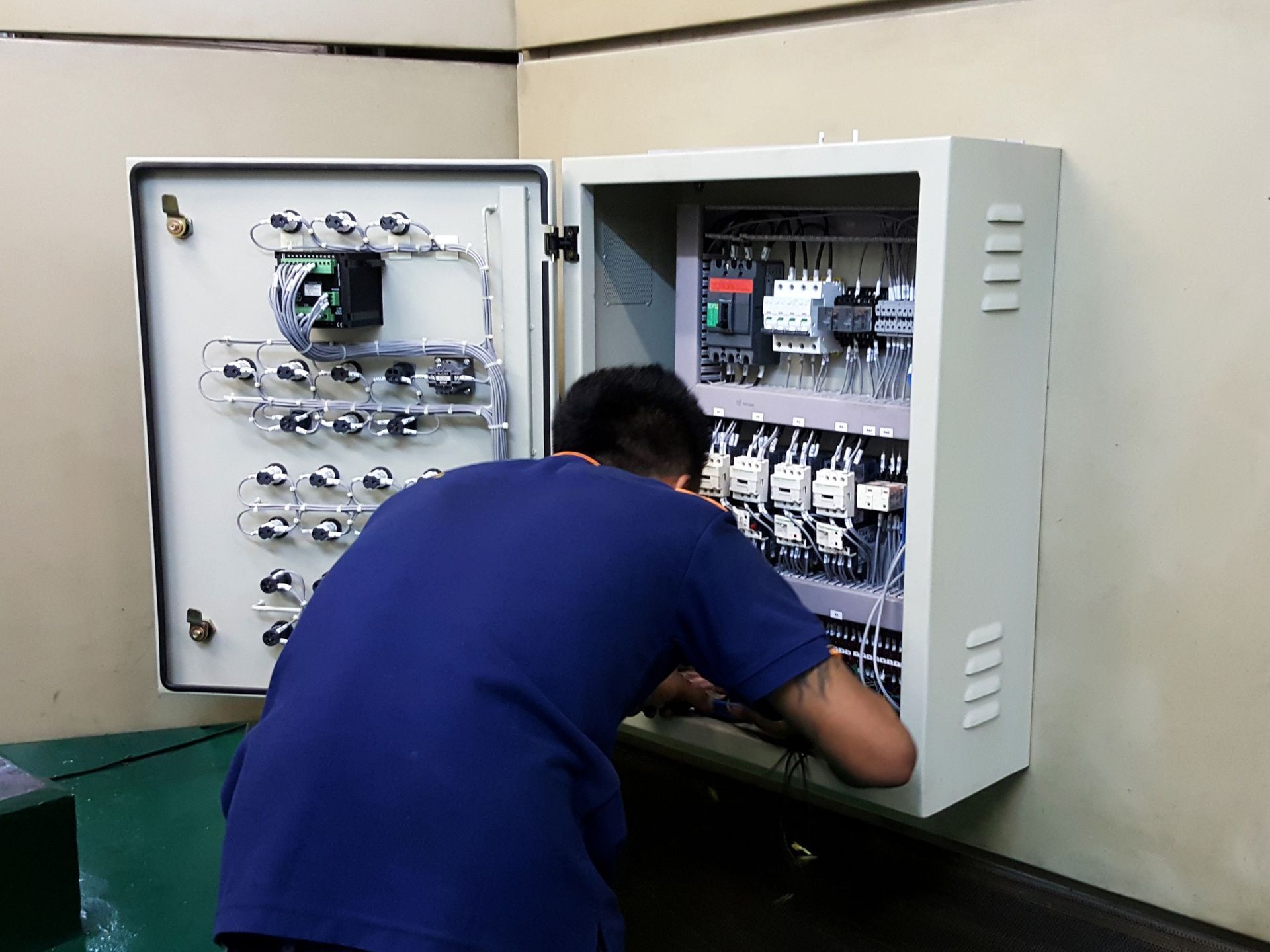 Person working on an open electrical control panel mounted on a beige wall.