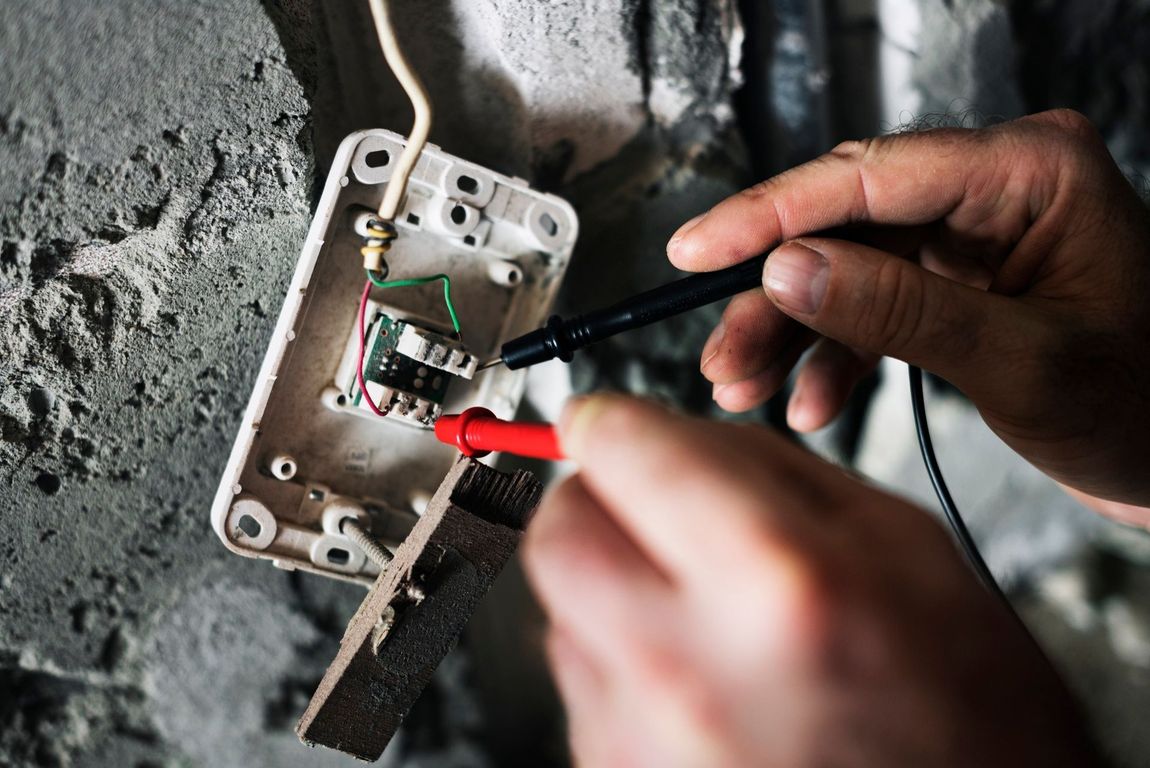 Hands testing wires in an open electrical outlet on a gray wall with a black and a red probe.