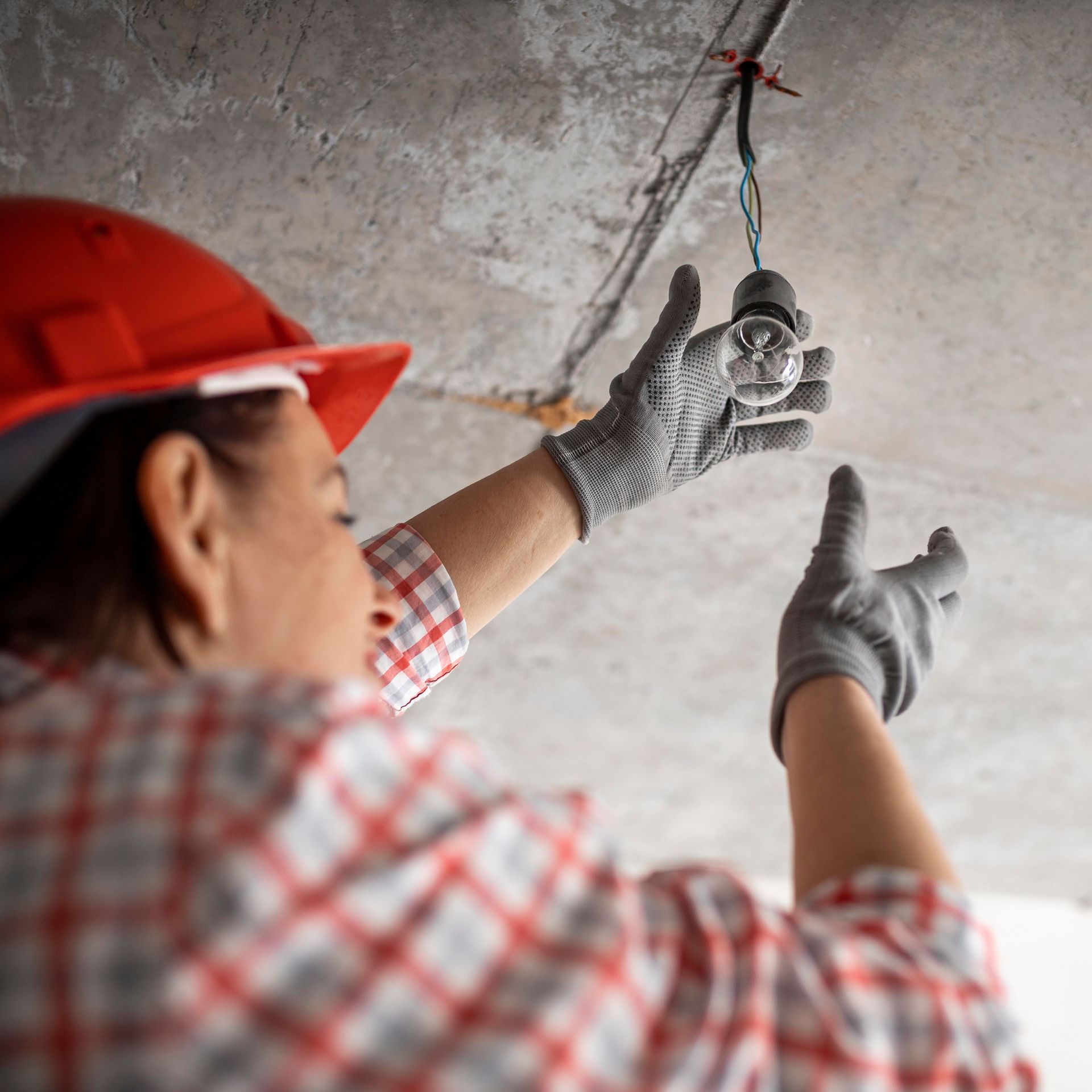 Person wearing red hard hat and gloves working on a light fixture attached to a concrete ceiling.