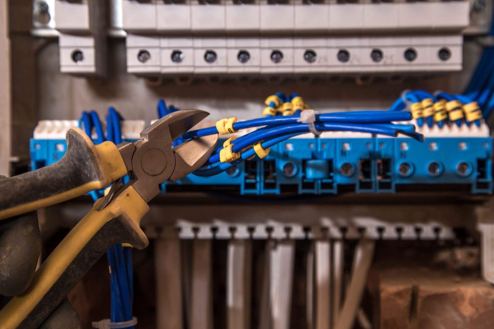 Person using wire cutters on blue electrical wires in a panel with circuit breakers and terminals.