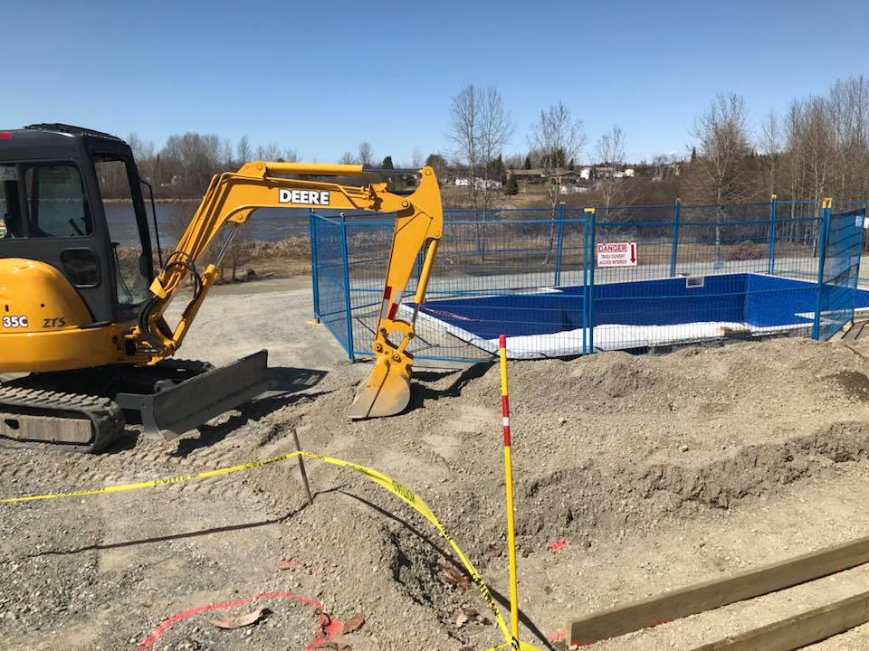 Excavatrice jaune creusant un terrain près d'une zone clôturée avec barrières bleues.