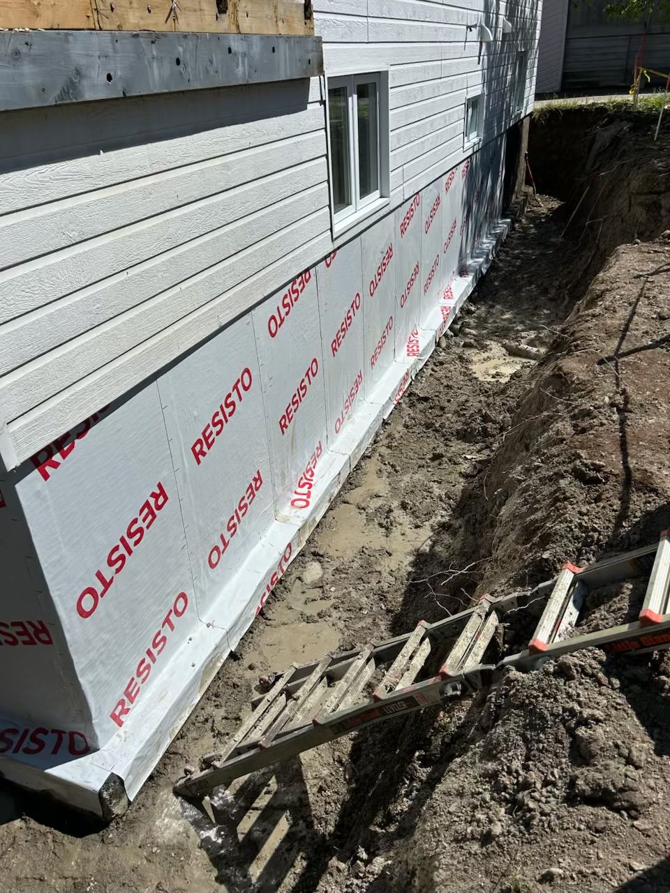 Fondation de maison avec membrane blanche de protection et tranchée de drainage excavée.