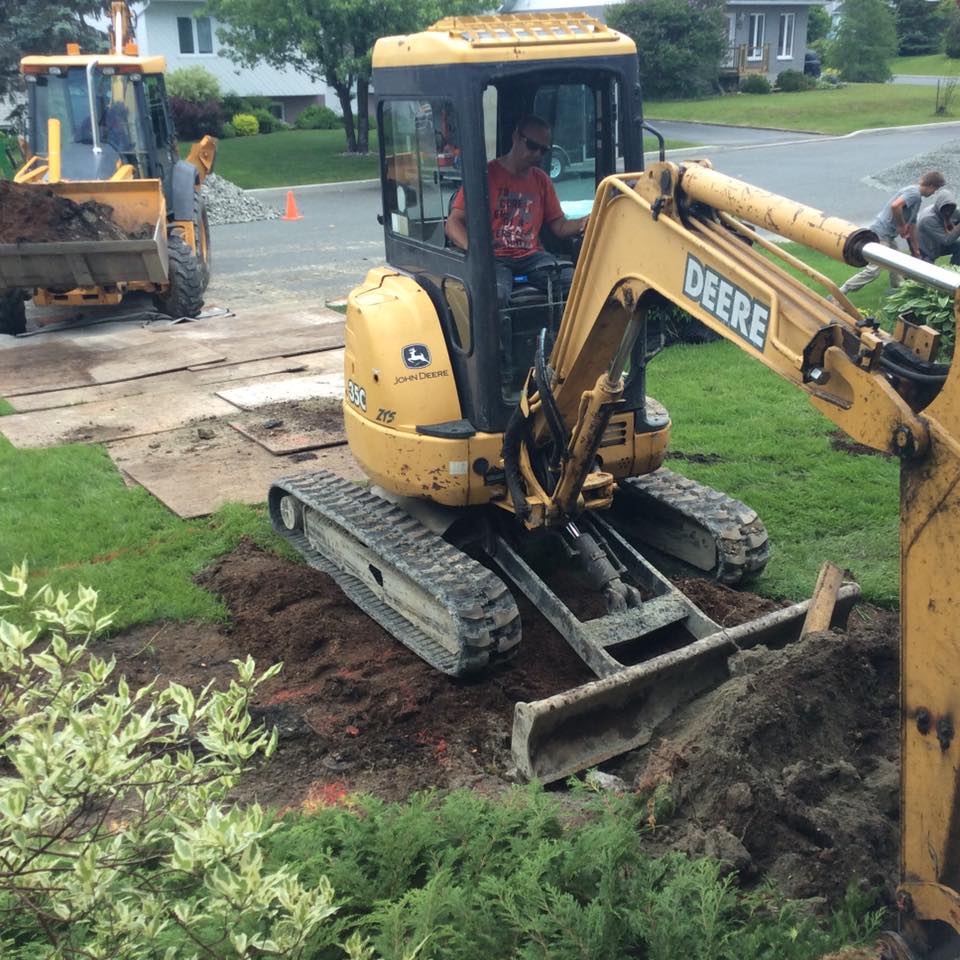 Mini excavatrice jaune creusant le sol d'une cour résidentielle avec d'autres équipements de construction visibles.