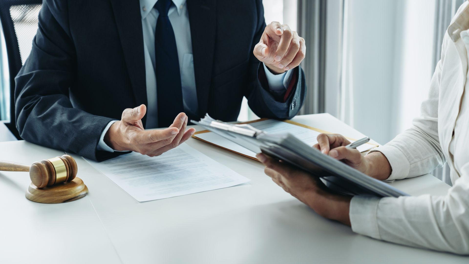 Man in suit gestures, discussing documents with person holding tablet, gavel on desk.
