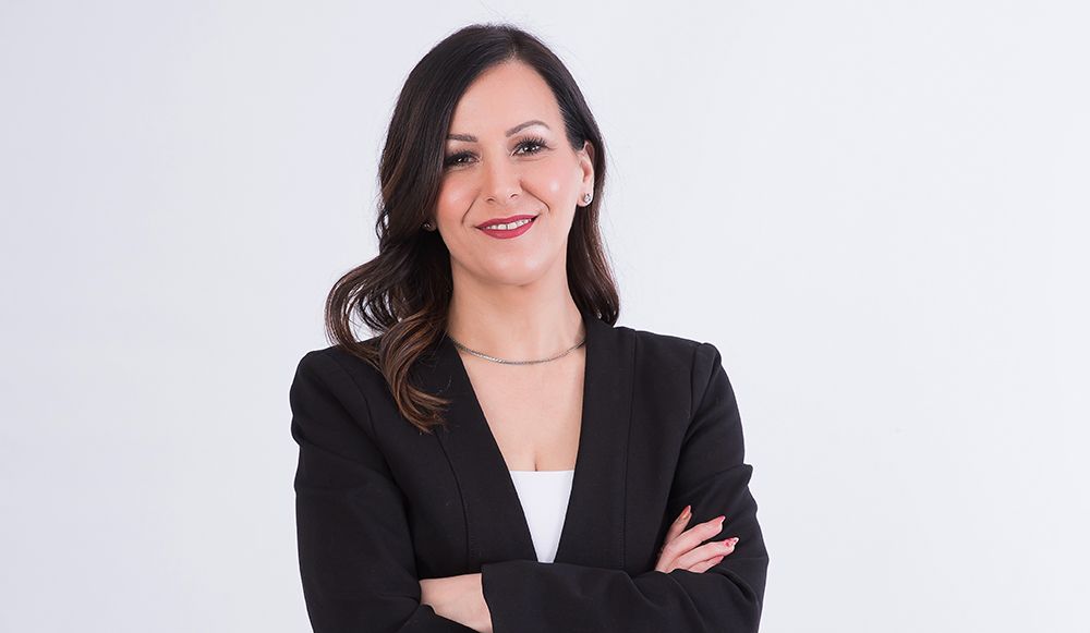 Woman with dark hair, wearing a black blazer and white top, arms crossed, smiling.