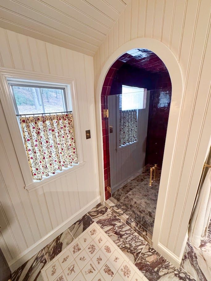Narrow hallway with white paneling, patterned wallpaper, arched doorway, and small windows with blinds