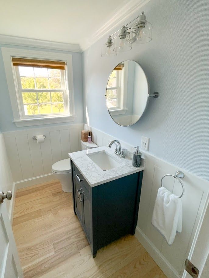 Bright small bathroom with white walls, wood floor, vanity, mirror, toilet, and window light.