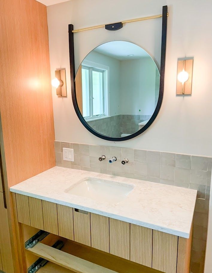 Modern bathroom vanity with round mirror, white countertop, wall sconces, and light wood cabinet