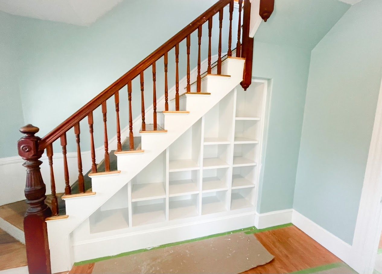 Staircase with white built-in storage shelves beneath, white steps, and wooden banister in a light blue room
