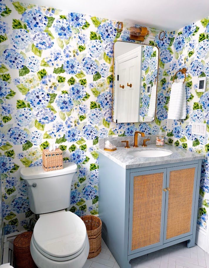 Floral wallpaper bathroom with white toilet, blue vanity, gold faucet, and woven cabinet doors