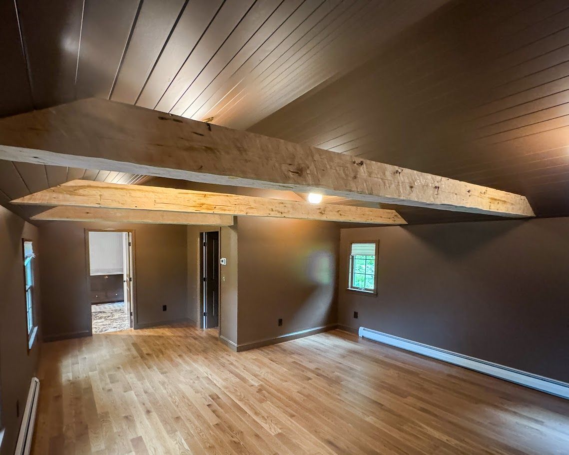 Empty attic room with wood floor, sloped ceiling, exposed beam, and small window