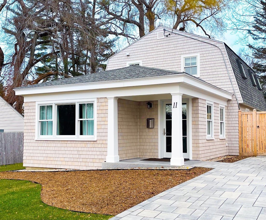 Small beige house with a gray roof, white trim, and a paved walkway in front