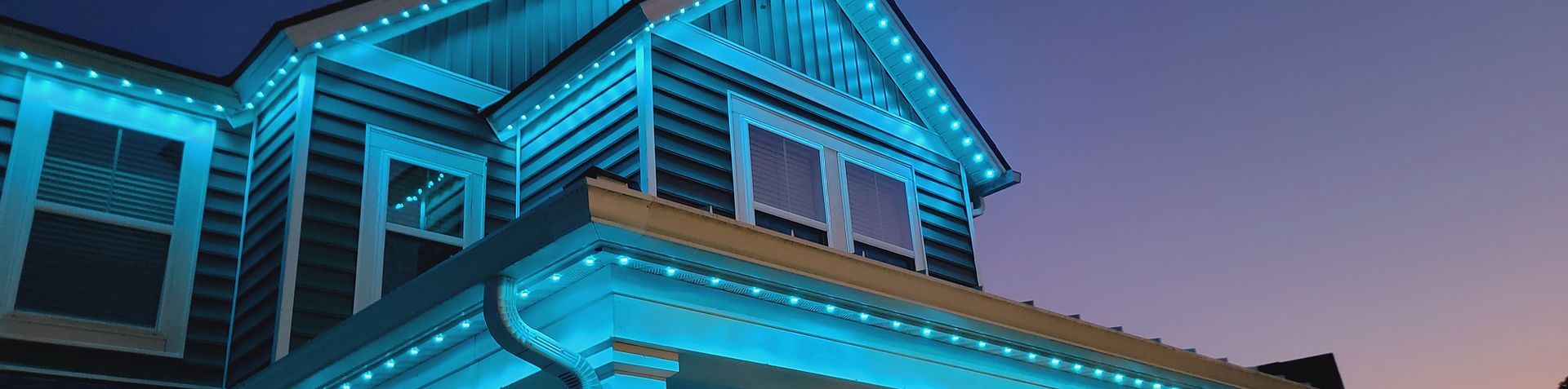 Blue-lit house exterior at dusk, showing the roofline and porch with warm interior lights.
