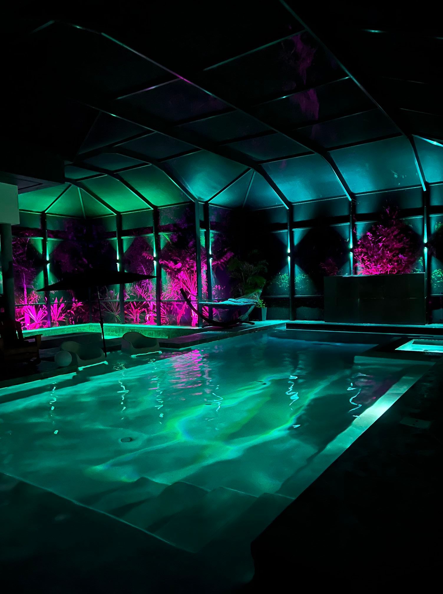 Neon-lit indoor pool with turquoise water, magenta plants, and a dark glass ceiling