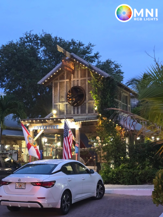 White sedan parked outside a lit, rustic building at dusk with flags and greenery.