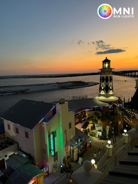 Sunset over a seaside resort with a lit tower, pool, and buildings beside the beach