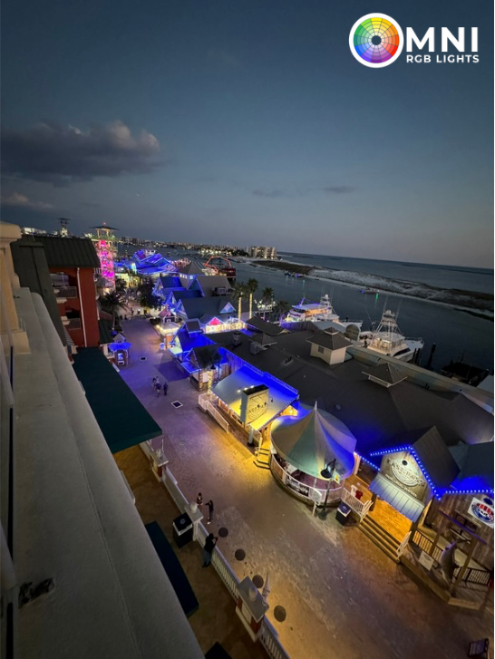 Nighttime harbor with yachts, blue-purple lights, and a waterfront road beside the sea