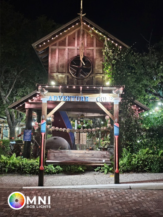 Nighttime view of a wooden gazebo-like structure with lights and greenery, labeled MNI at the bottom.
