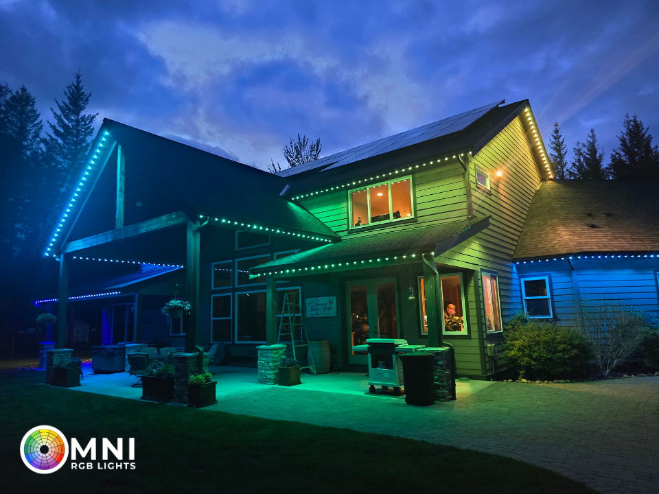 Night-lit modern house with blue and green accent lights, deck seating, and a glowing window at dusk