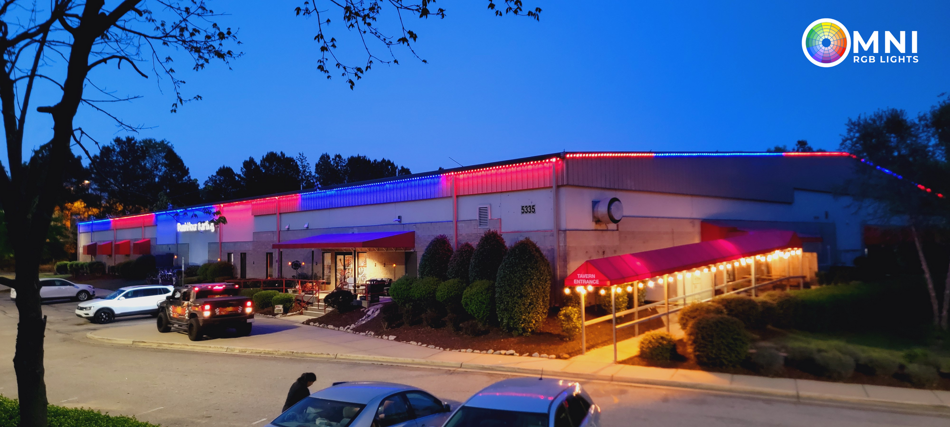 Twilight view of a hospital building lit in red, white, and blue with parked cars in front