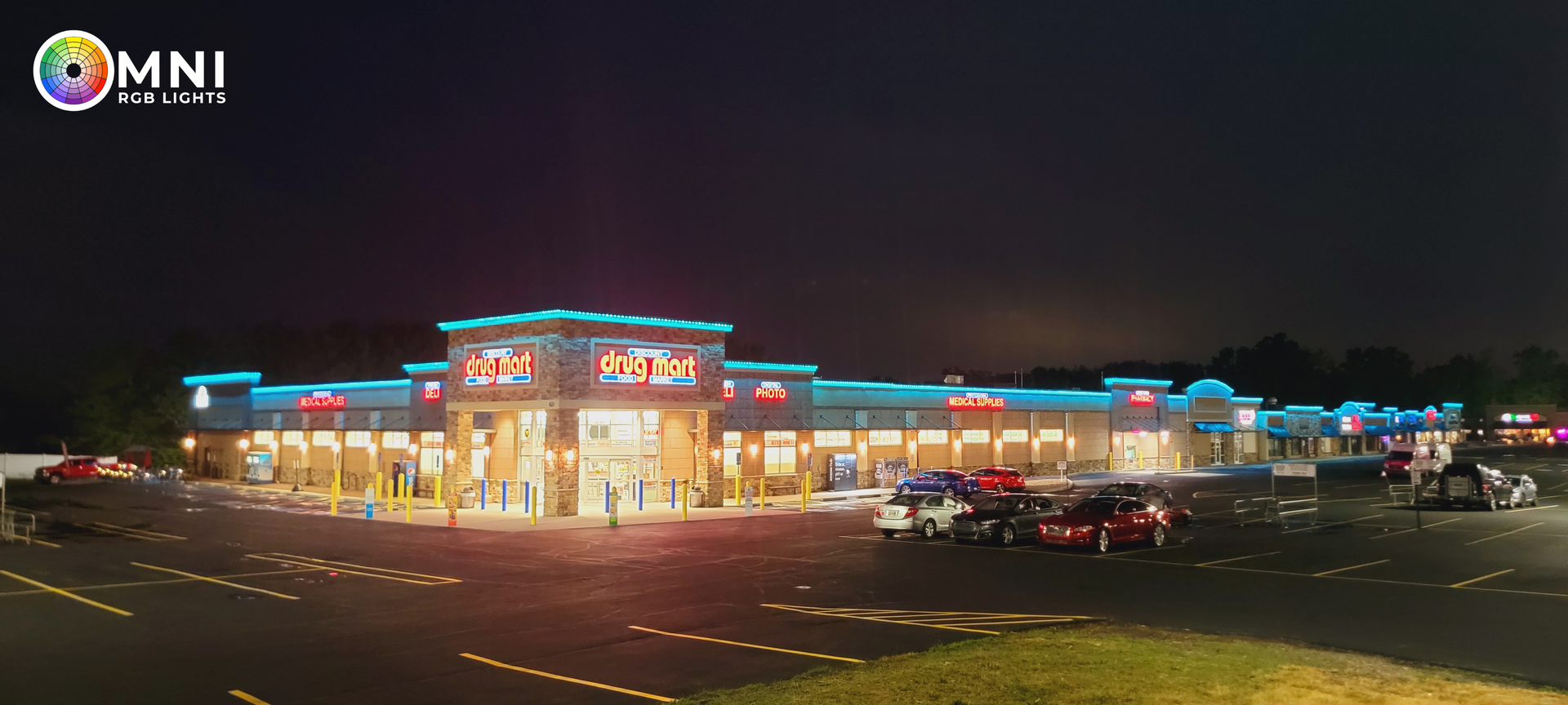 Nighttime view of an illuminated shopping plaza with parking lot and cars under a dark sky