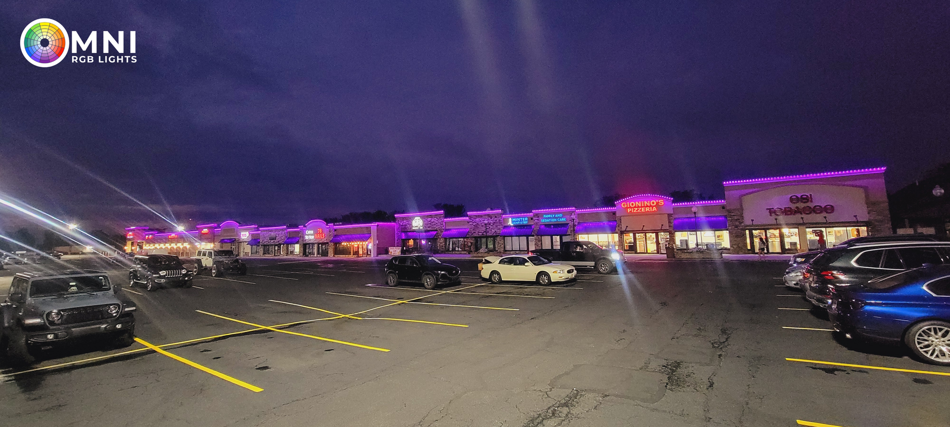 Nighttime parking lot with many parked cars and a long line of purple and pink lit tents or buildings