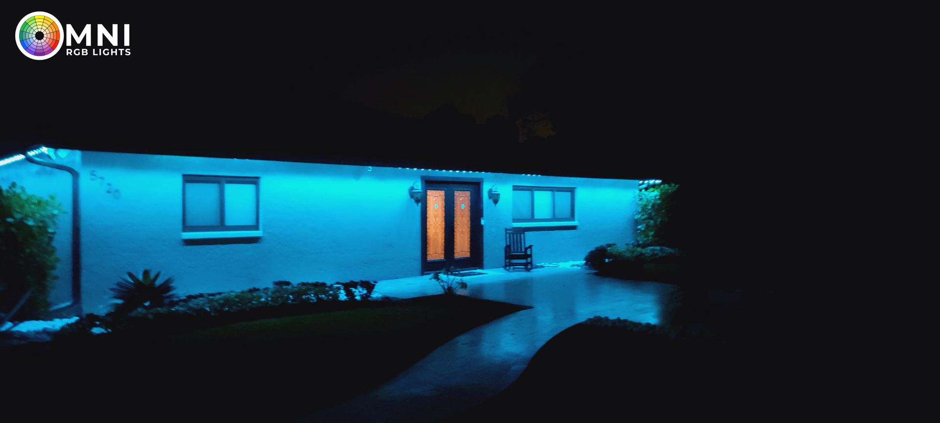 Blue-lit modern house at night with illuminated doorway, windows, and a dark walkway leading to the entrance