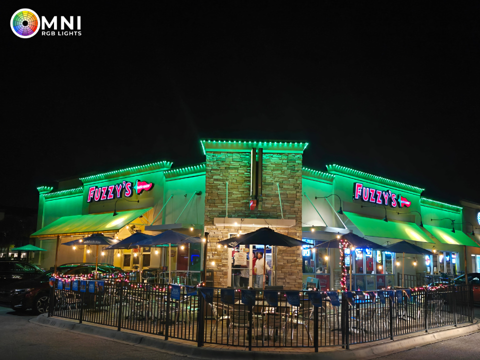 Nighttime exterior of a brightly lit restaurant with green neon accents and patio seating behind a fence.