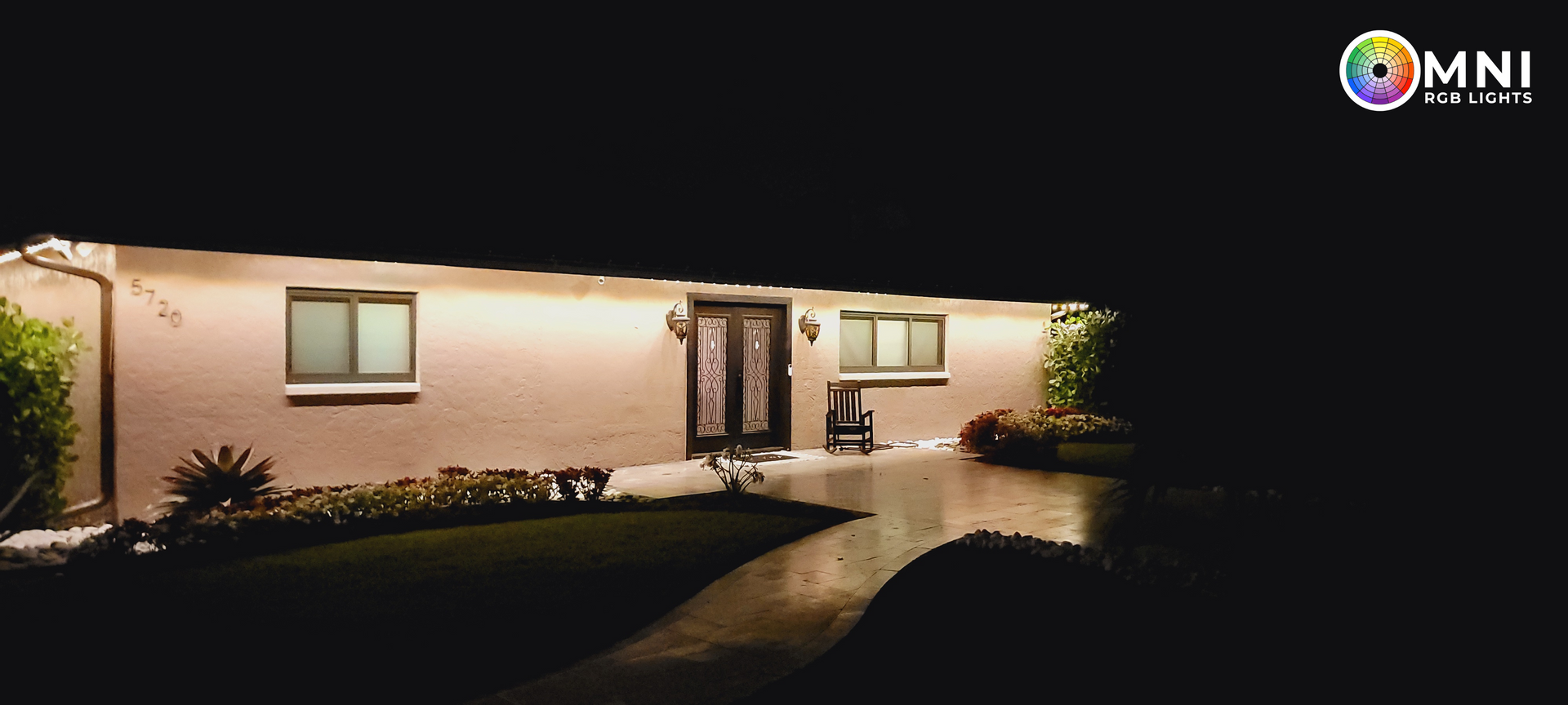 Small white house lit at night with a front porch, windows, and a wet walkway leading to the entrance