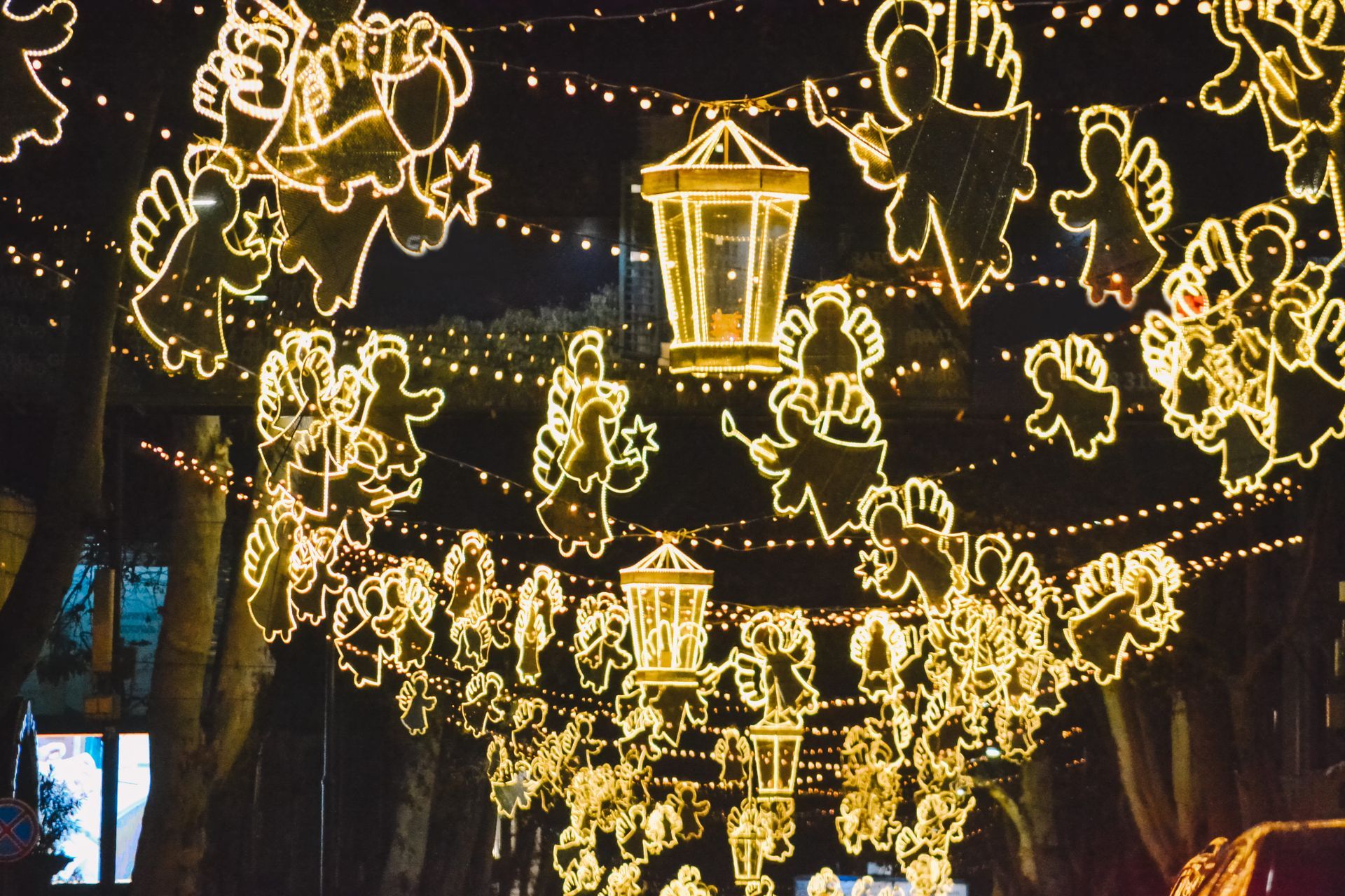 Glowing holiday lights and angel shapes hanging above a street at night, with a lantern centerpiece