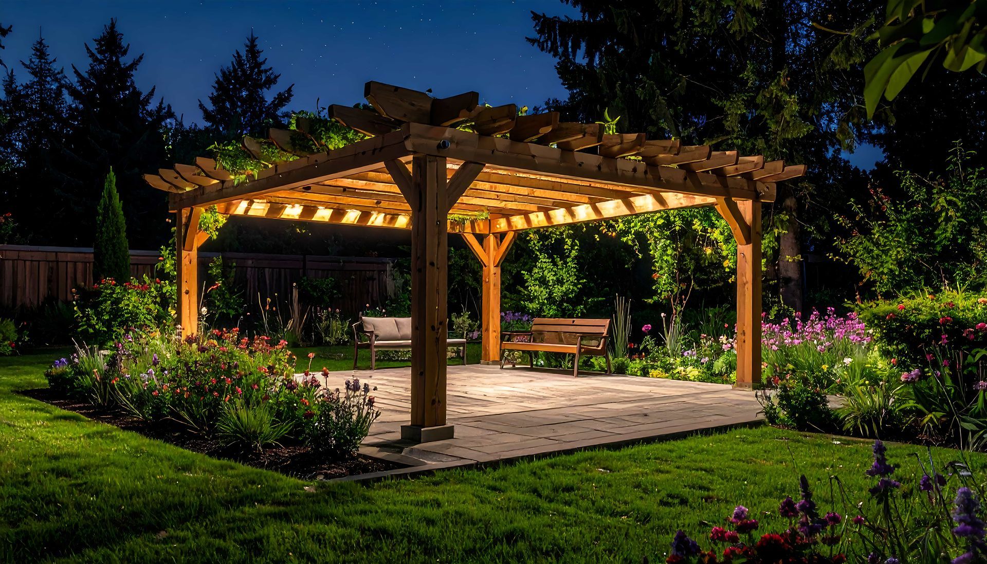 Illuminated wooden garden pergola at night, surrounded by flowers and trees, with stone flooring and warm lights