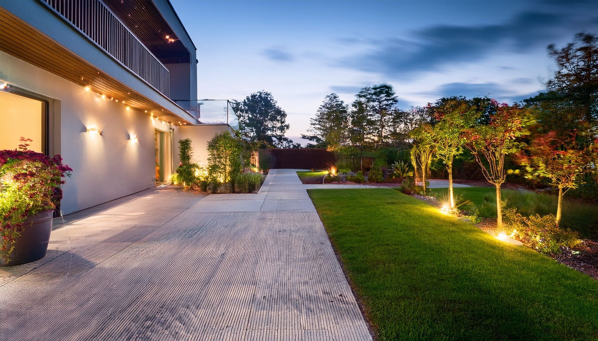 Modern home exterior with lit garden, stone walkway, and manicured lawn at dusk