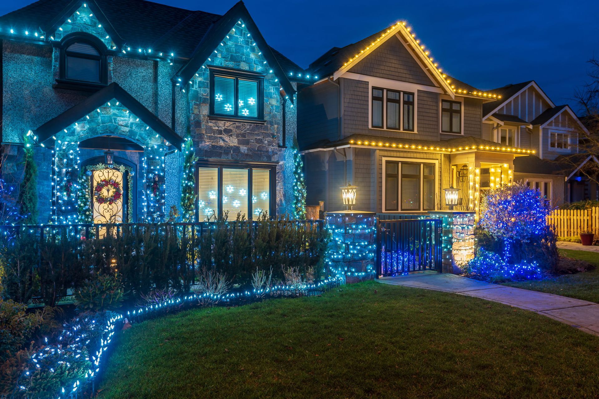 Suburban homes lit with blue and gold holiday lights at night, with a glowing yard and walkway