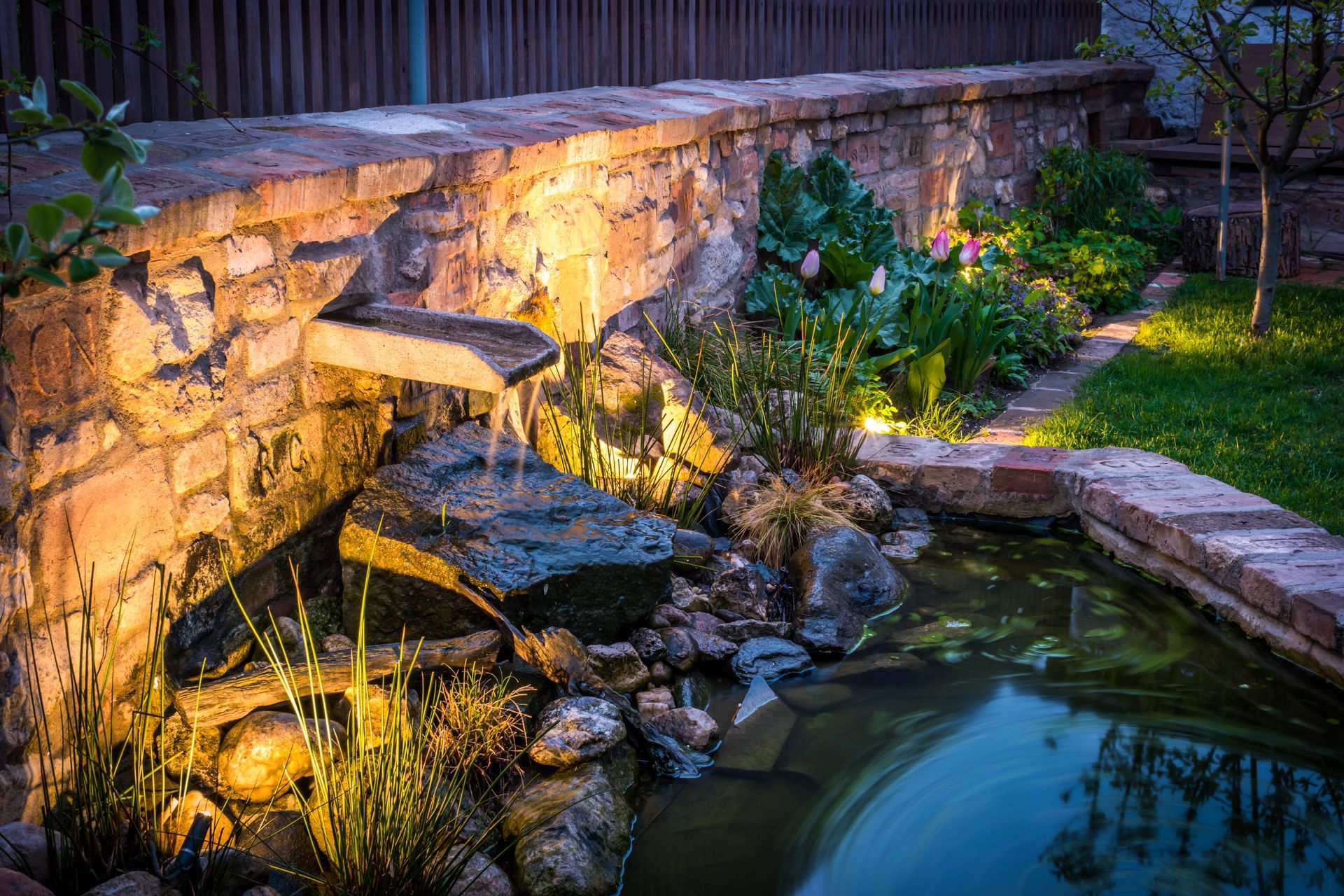 Lit stone garden waterfall flowing into a pond at dusk, with glowing lights and greenery