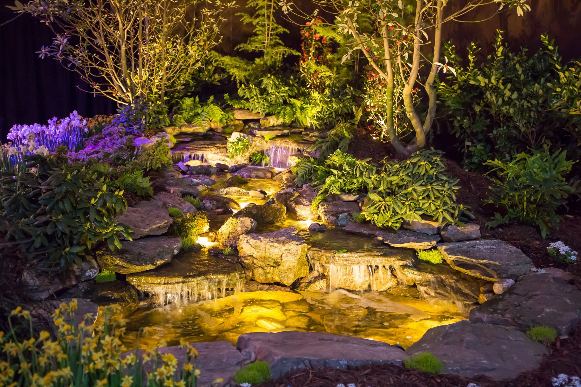 Illuminated garden waterfall over rocks, surrounded by lush greenery and purple flowers at night