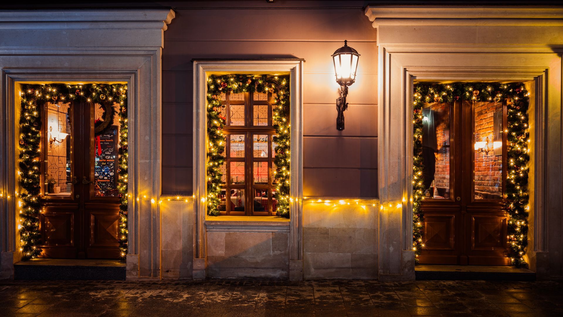 Three lit windows and a door decorated with warm holiday lights on a building exterior at night