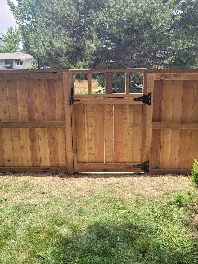 Wooden gate in a cedar fence, set in a grassy yard, with a treeline visible in the background.