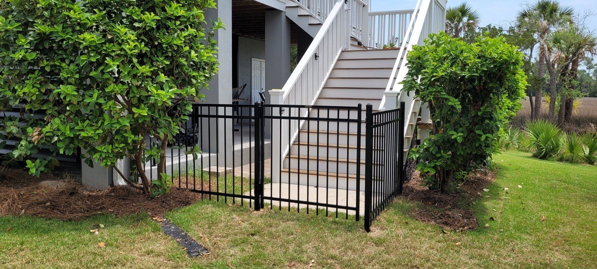 Black metal fence in front of stairs leading to a building, flanked by green bushes and grass.