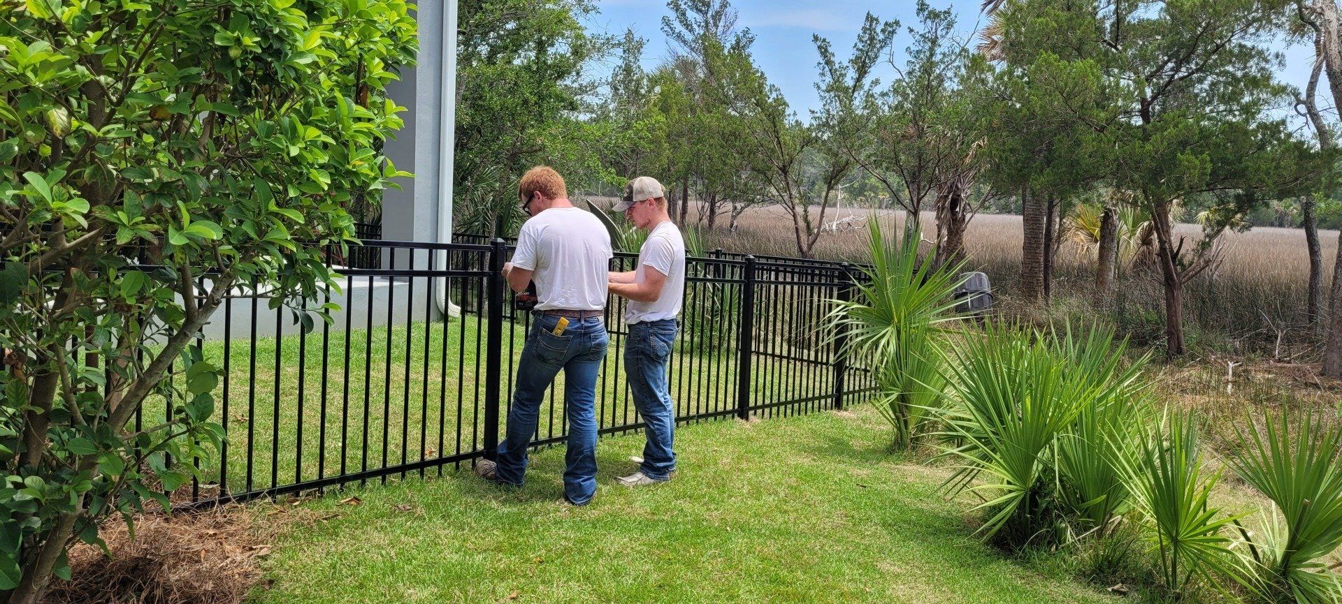 Two people install a black metal fence in a yard with green grass and trees.