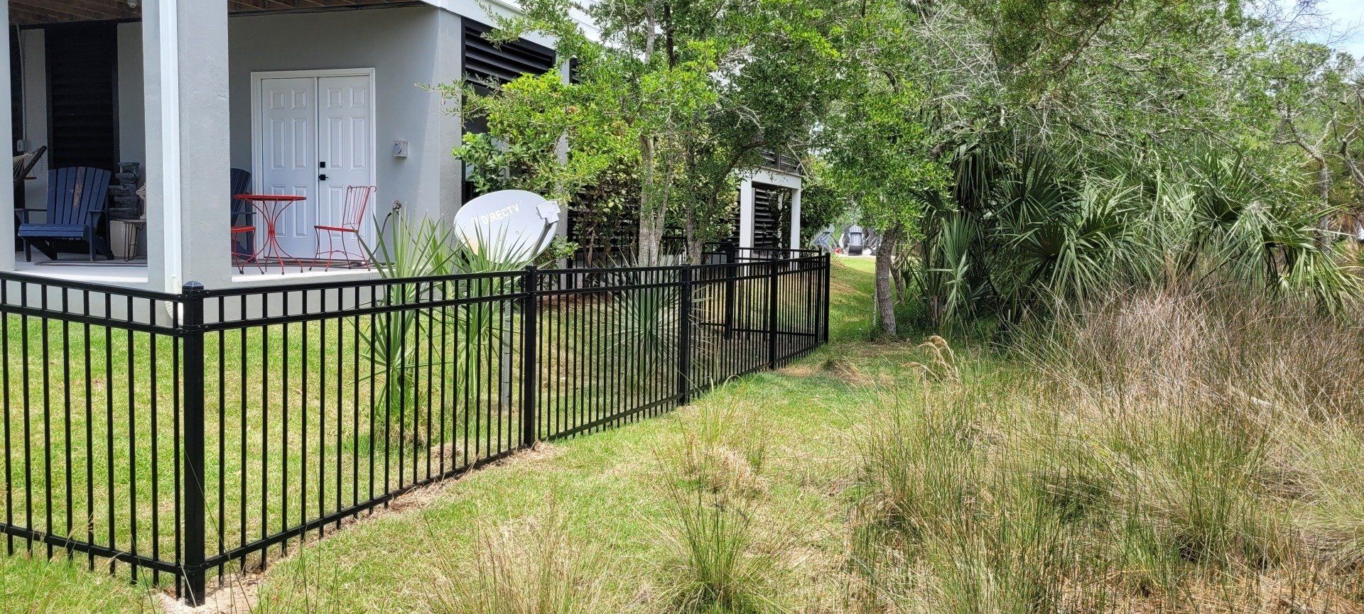 A black metal fence surrounds a house with a white door and satellite dish, with overgrown grass on the side.
