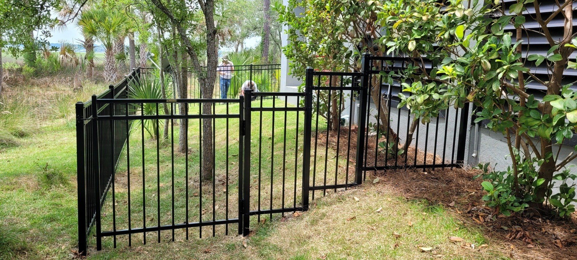 Black metal fence section in a yard with trees and bushes.