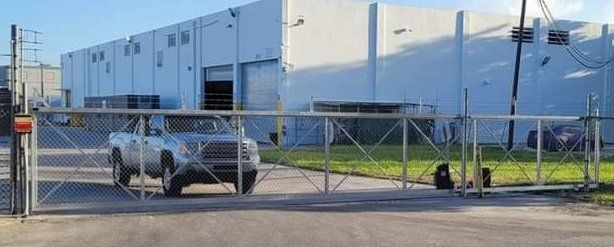 Silver pickup truck at a gate leading to a large building.