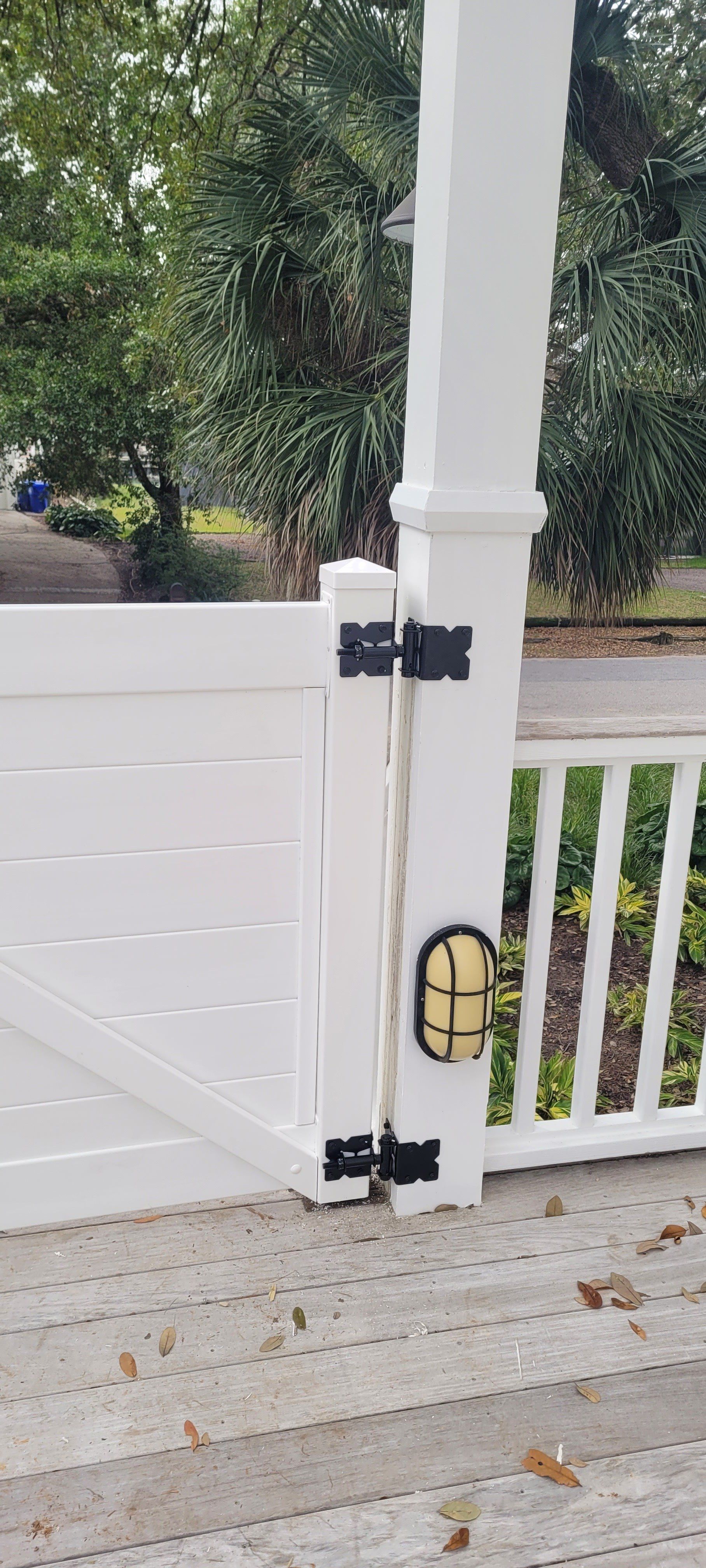 White porch gate with black hinges, light fixture on post, and greenery in the background.