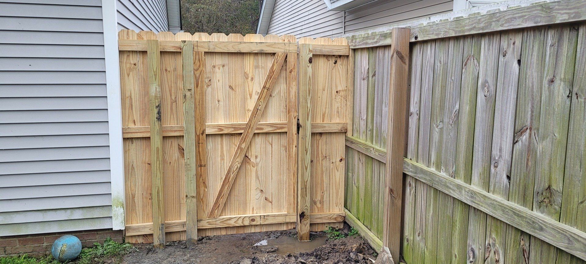 Wooden fence gate with diagonal brace, between two wooden fences, next to a building.