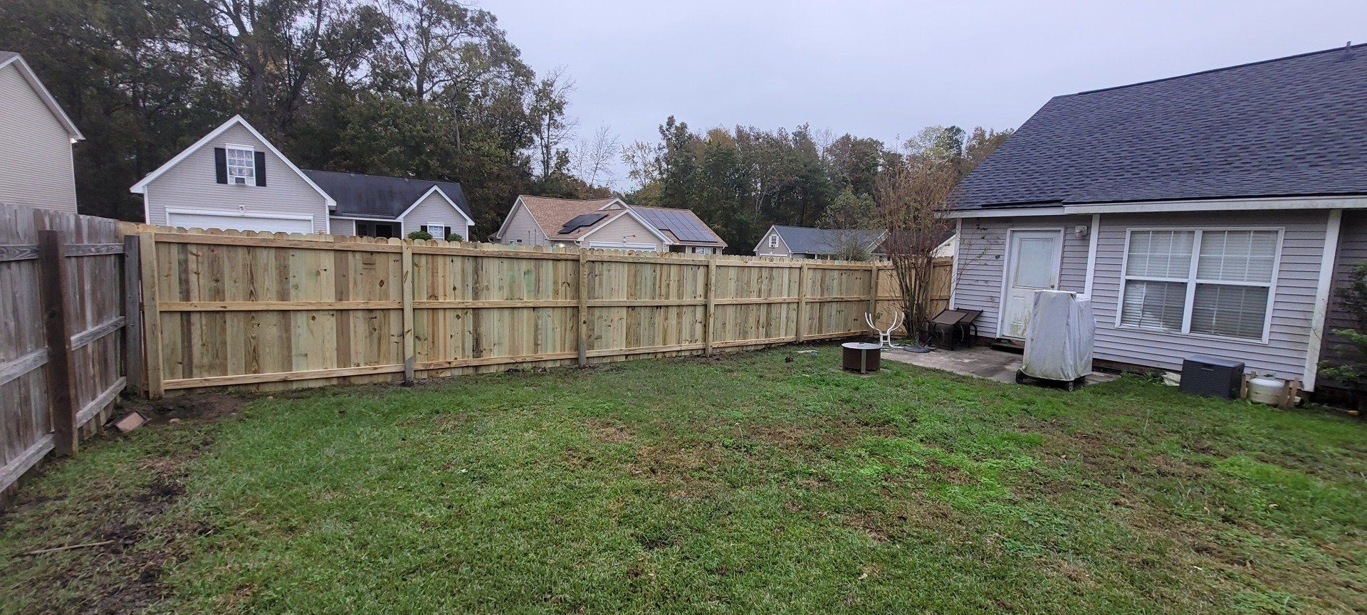 Backyard with a wooden fence. Houses and trees are in the background. Overcast sky.