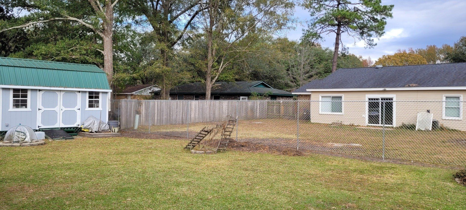 Backyard with shed, fence, and house under cloudy sky. Green grass, trees.