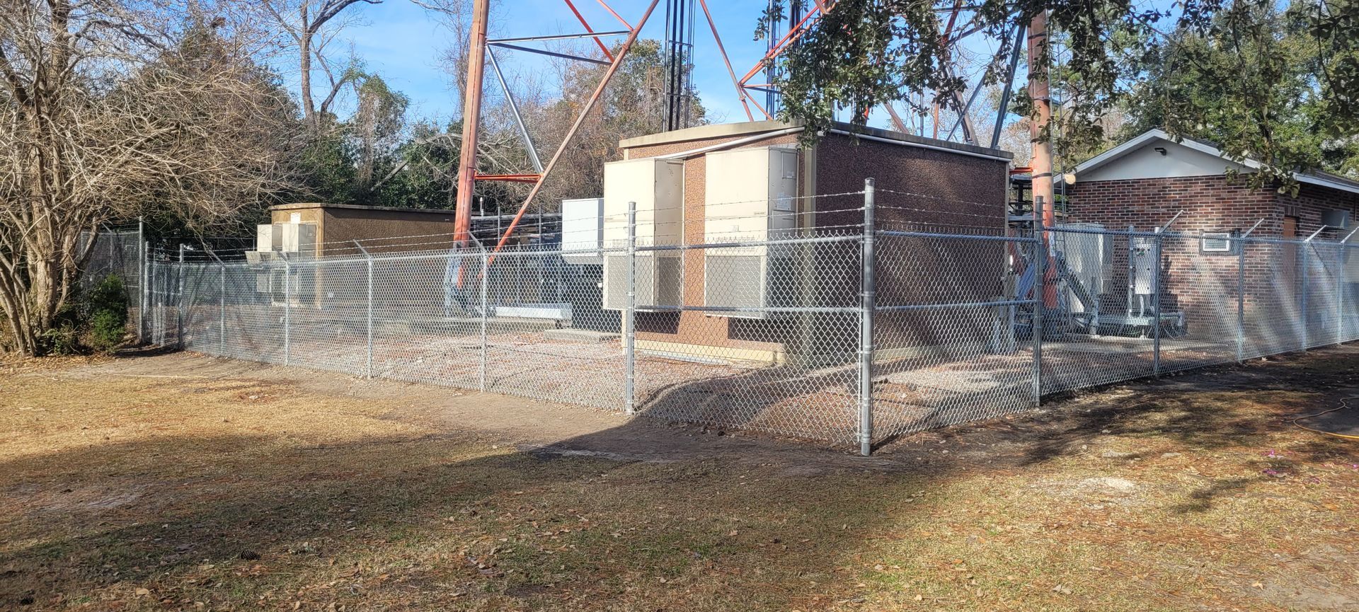 A fenced industrial site with a radio tower and beige buildings. Brown grass is in the foreground.