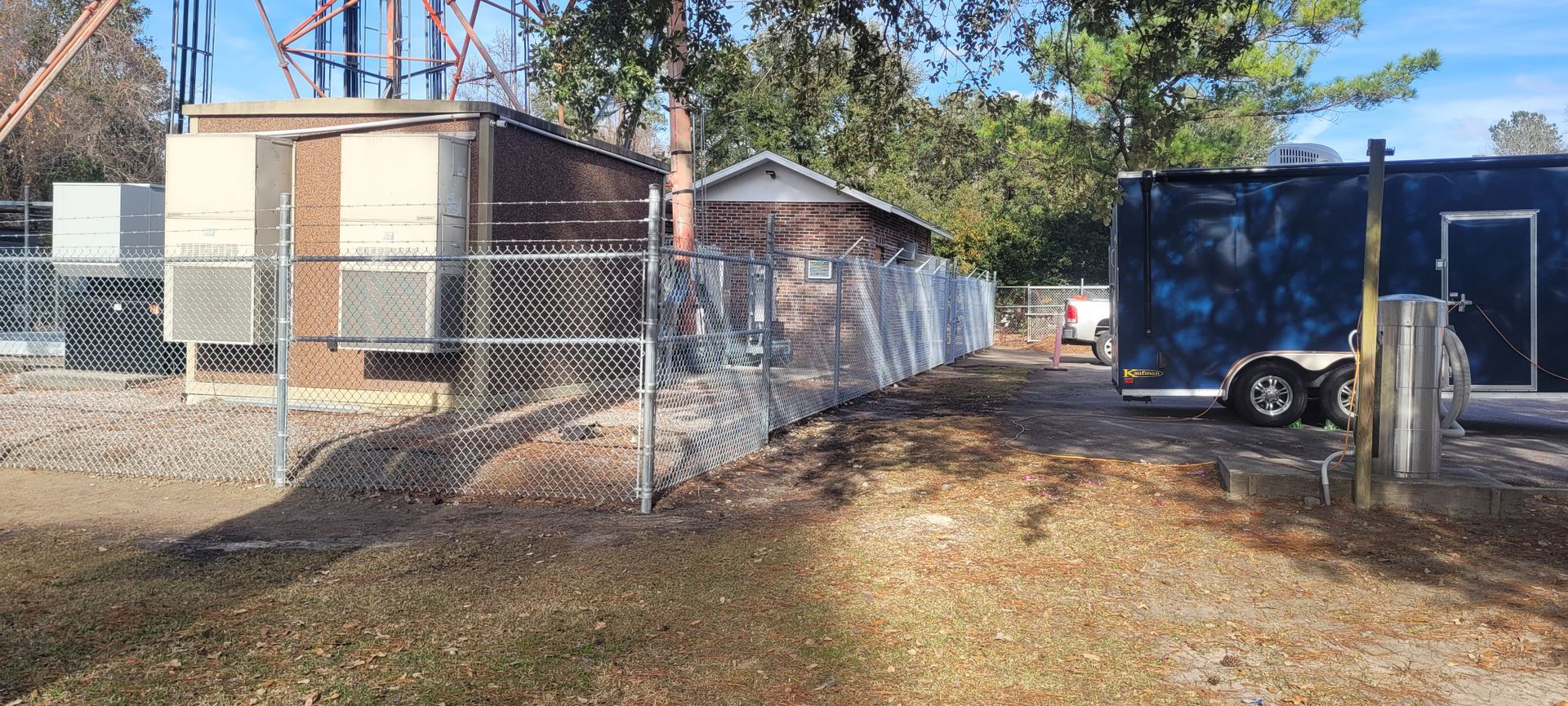 Fenced industrial area with a trailer, buildings, and a glimpse of a vehicle.