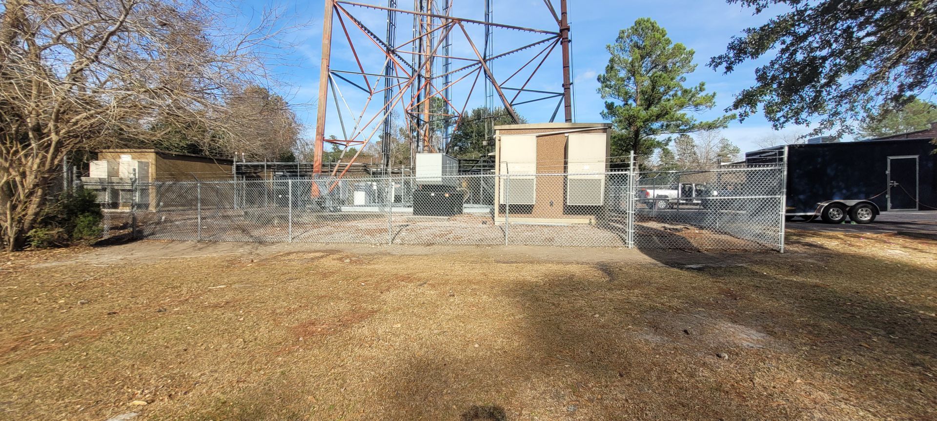 A fenced-in utility site with a metal tower, shed, and trailer on a sunny day.