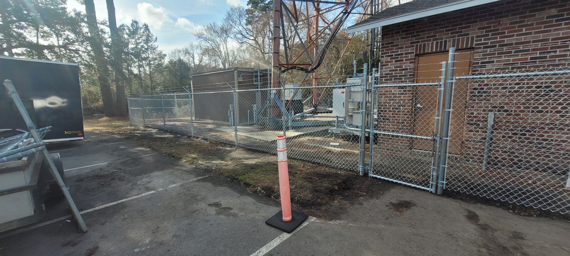 A fenced-in utility area with a brick building, a traffic cone, and a telecommunications tower.